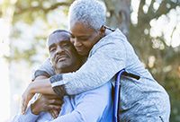 Photo of an elderly couple. The husband is in a wheelchair, the wife is hugging him.